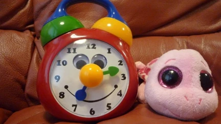 A colourful toy clock with a smiling face sitting on a sofa next to a pink toy turtle