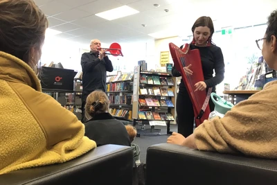 Framed between two audience members, a harpist playing a red harp leans towards them while in the background is a trombonist playing a red trombone