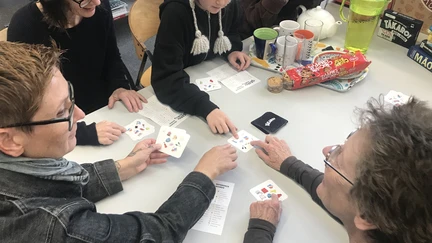 A small group of adults, children and teens are sitting down together at a table, playing a board game in te reo Māori. There are cups of tea and packets of biscuits in the background.