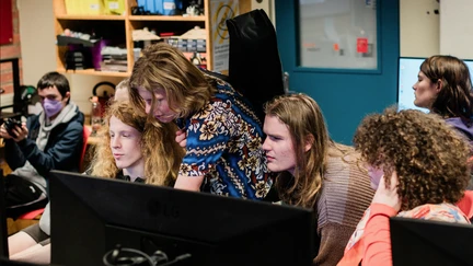 A group of rangatahi are clustered around a computer as a tutor shows them something on the screen.